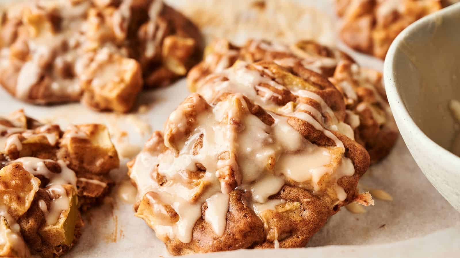 Close-up of freshly baked apple cookies drizzled with icing, resting on parchment paper with part of a bowl visible to the right.