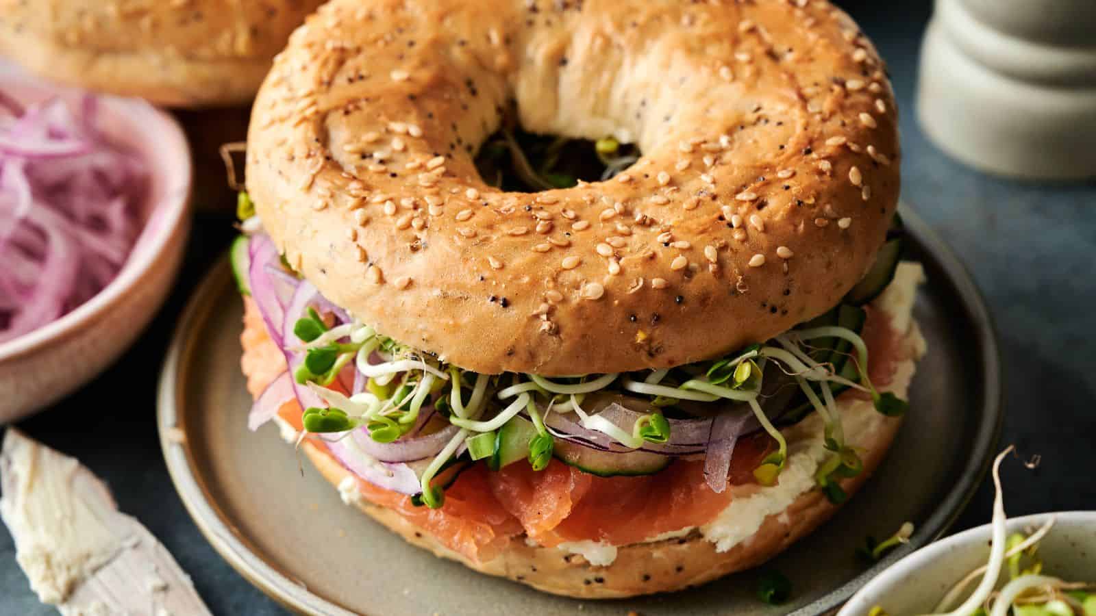 A sesame seed bagel sandwich with cream cheese, smoked salmon, red onion, cucumber, and sprouts on a plate.