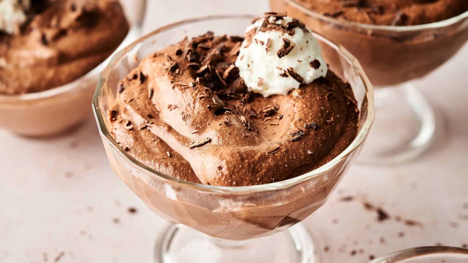 A glass dessert cup filled with chocolate mousse, topped with whipped cream and chocolate shavings, placed on a light-colored surface.