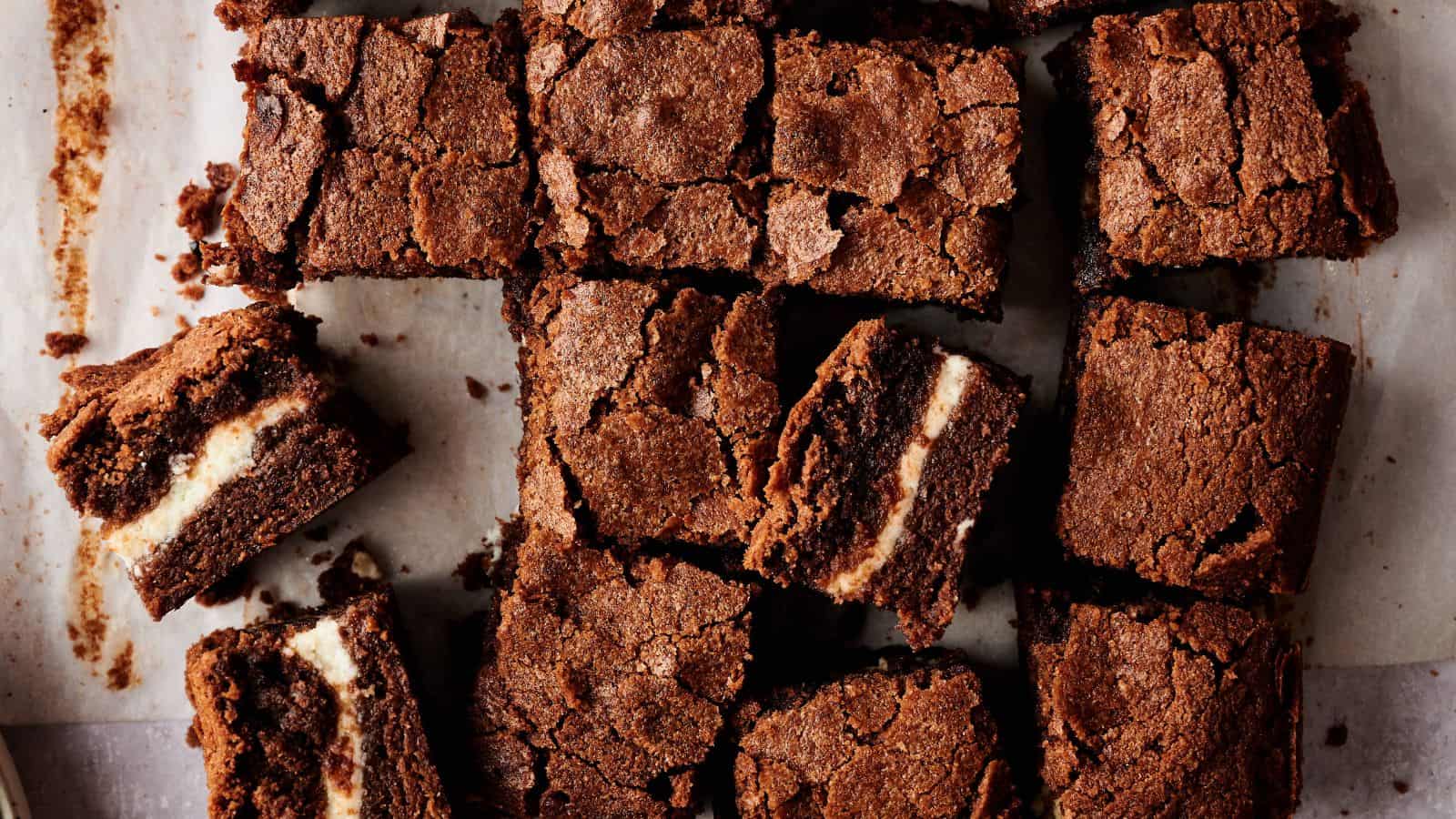Brownies with a cream cheese layer in the center are cut into squares and arranged on parchment paper.
