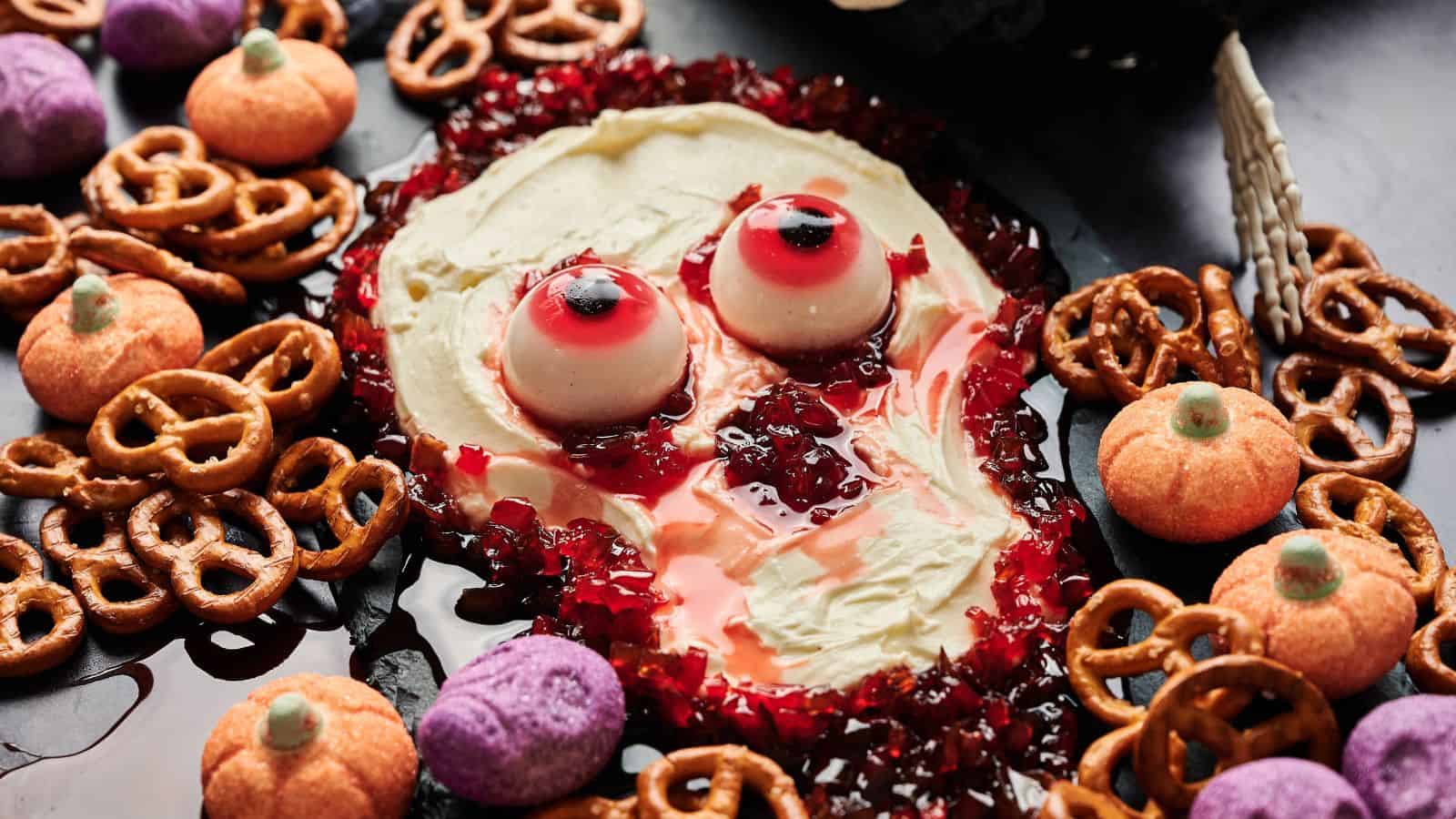 A Halloween-themed dip arranged to look like a spooky face with candy eyes, red jelly for blood, and surrounded by pretzels and festive snacks.