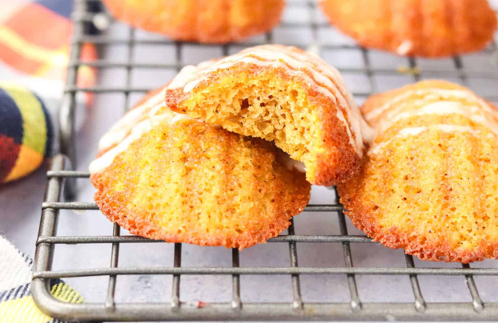 A madeleine baking tray holding several golden brown madeleine cookies, some drizzled with white icing, next to a cup of coffee and a checkered cloth.