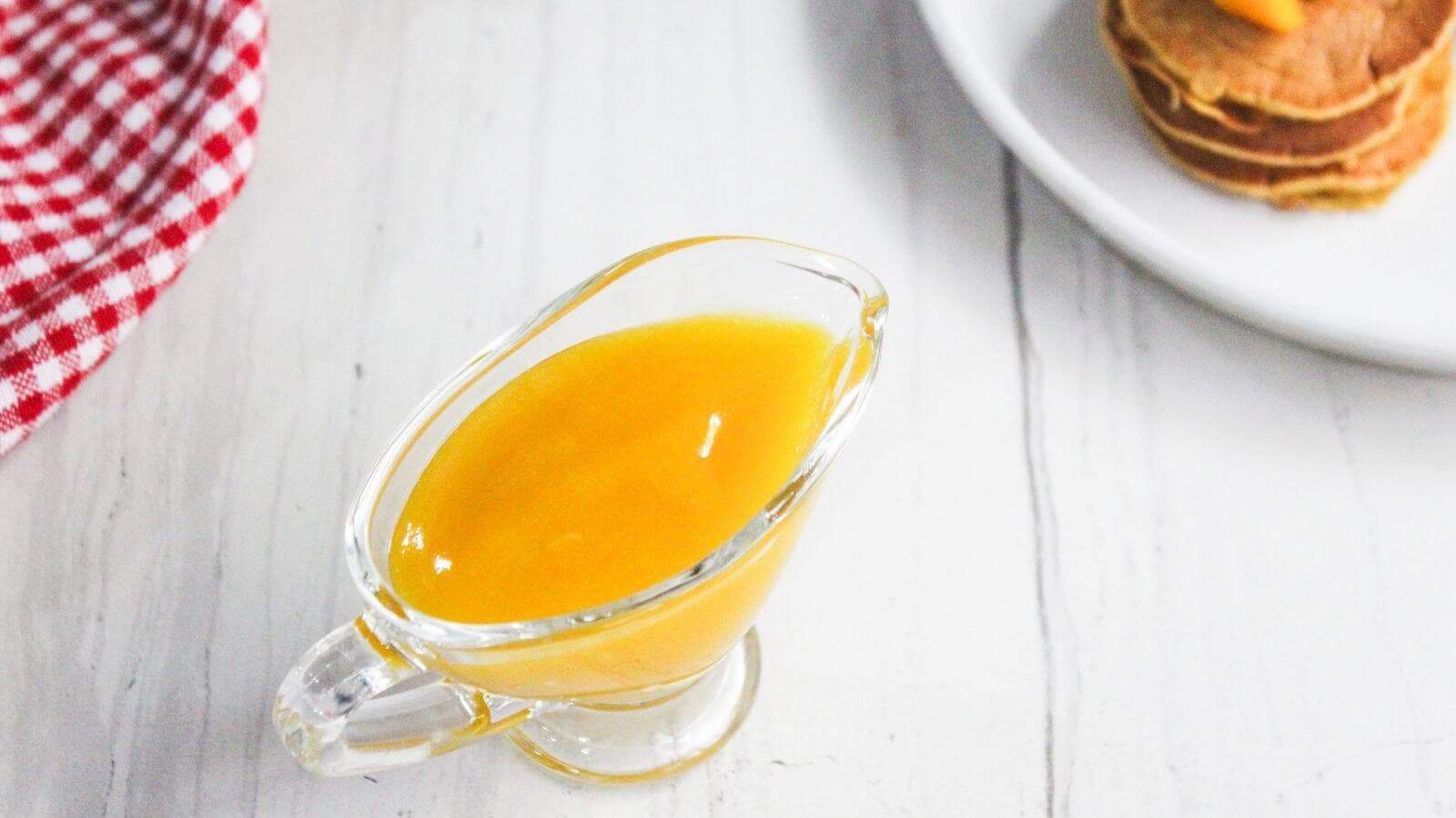 A glass gravy boat filled with mango sauce sits on a white surface next to a plate of pancakes topped with mango cubes and a red checkered cloth.