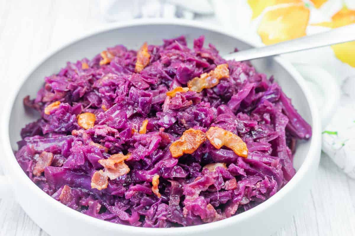 A bowl of cooked red cabbage mixed with small pieces of bacon, served with a spoon.