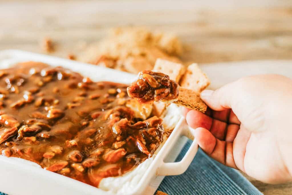 A hand dips a cracker into a casserole dish filled with a creamy white base topped with a chunky brown sauce.