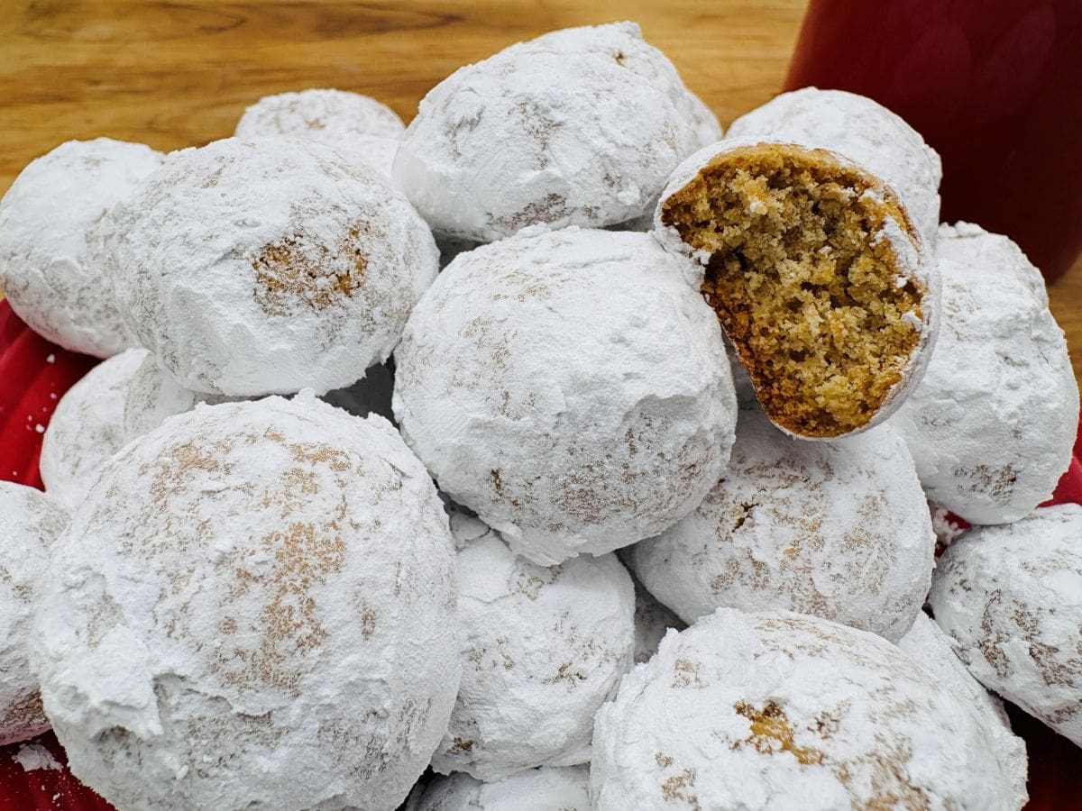 A pile of round cookies coated in powdered sugar, with one cookie broken open to show its crumbly interior.