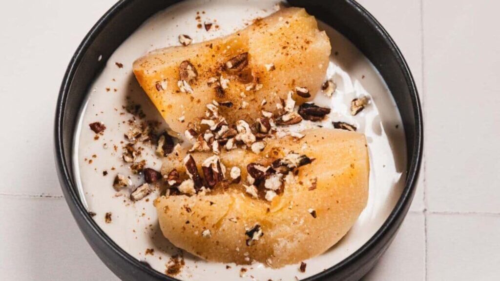 A bowl of yogurt topped with two peach slices, chopped nuts, and a sprinkle of cinnamon.