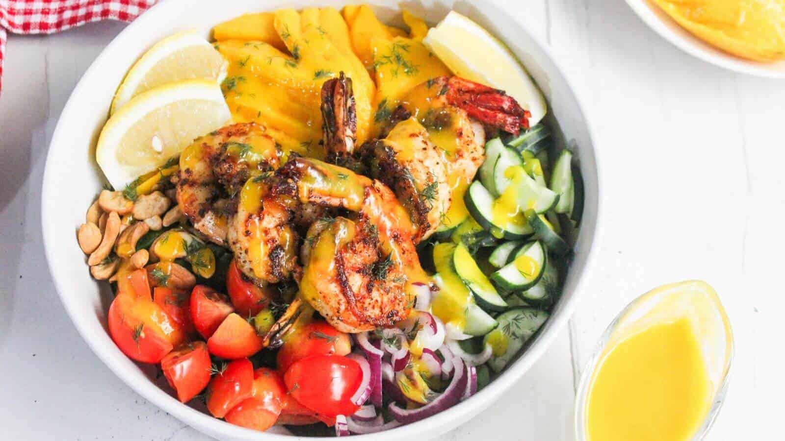 A bowl with grilled shrimp, sliced mango, lemon wedges, cherry tomatoes, cucumbers, red onions, peanuts, and greens, topped with a yellow sauce.