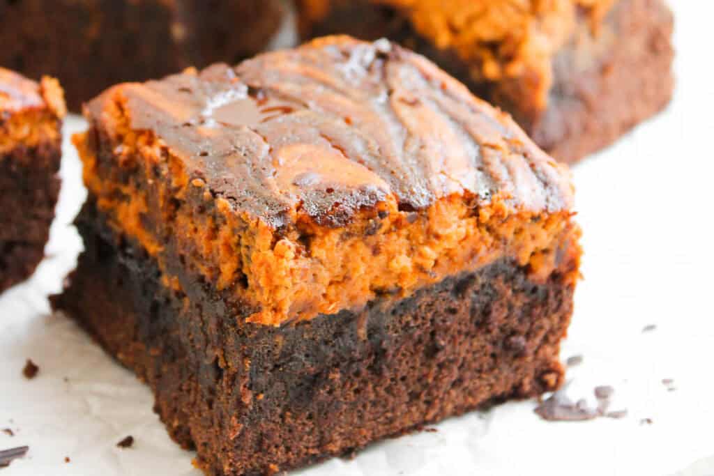 A close-up of a thick, square brownie with a marbled top layer, featuring a mix of dark chocolate and orange-brown swirls.
