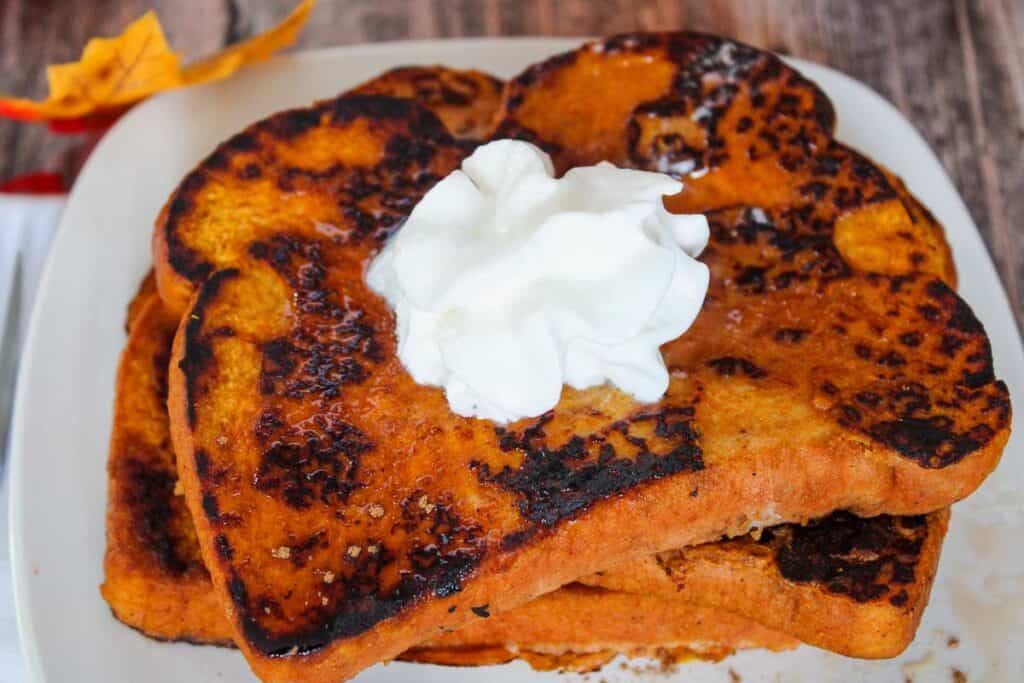 Two slices of pumpkin French toast stacked on a white plate, topped with a dollop of whipped cream.