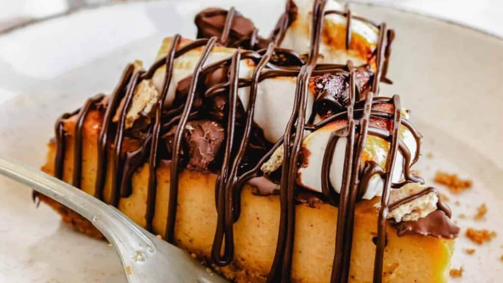 A slice of cheesecake topped with toasted marshmallows and chocolate drizzle on a plate with a fork.
