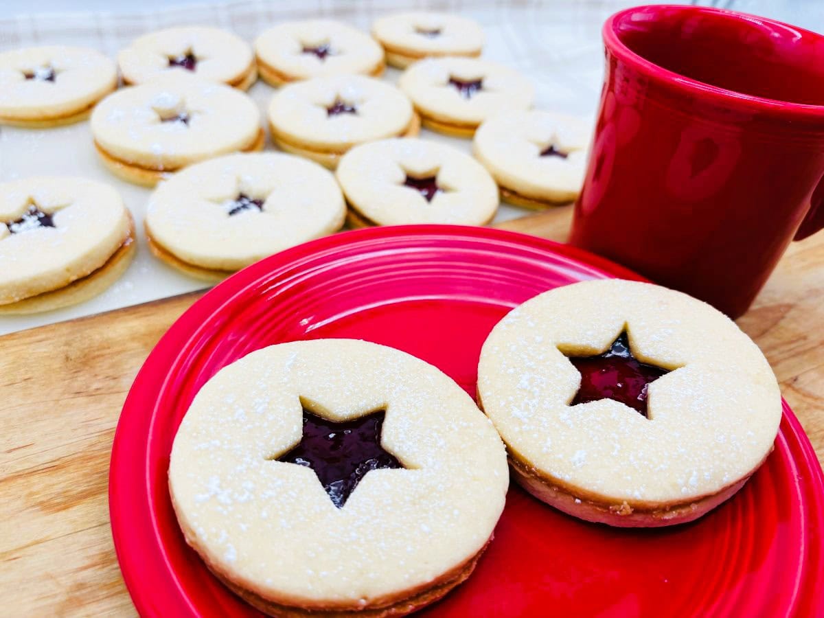 Two round sandwich cookies with star-shaped cutouts revealing jam, on a red plate next to a red mug. More cookies are on a tray in the background.