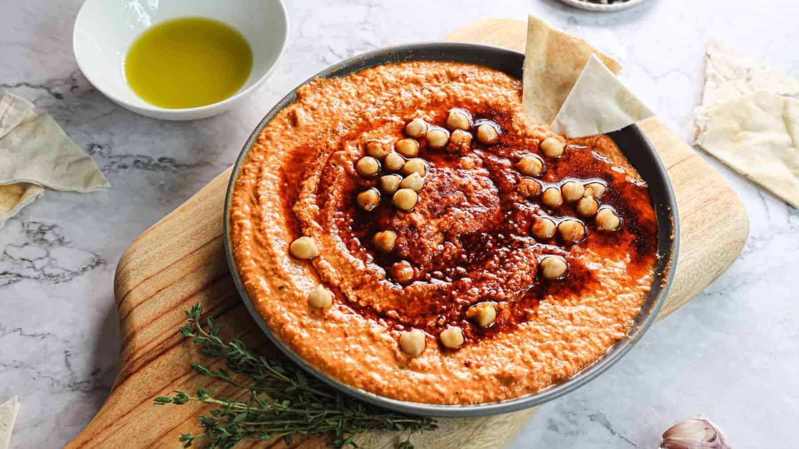 A bowl of creamy hummus topped with chickpeas and a red sauce, placed on a wooden board. Pita bread pieces and a small bowl of olive oil are nearby on a marble surface.