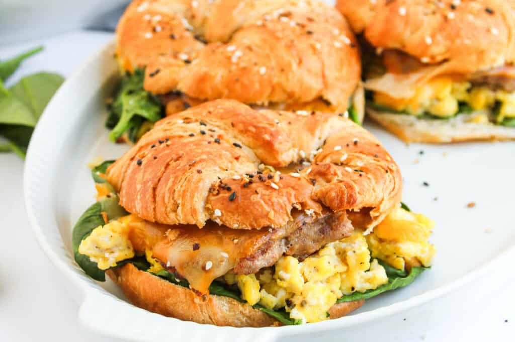 A croissant sandwich filled with scrambled eggs, cheddar cheese, spinach leaves, and sausage, garnished with seeds, served on a white plate.