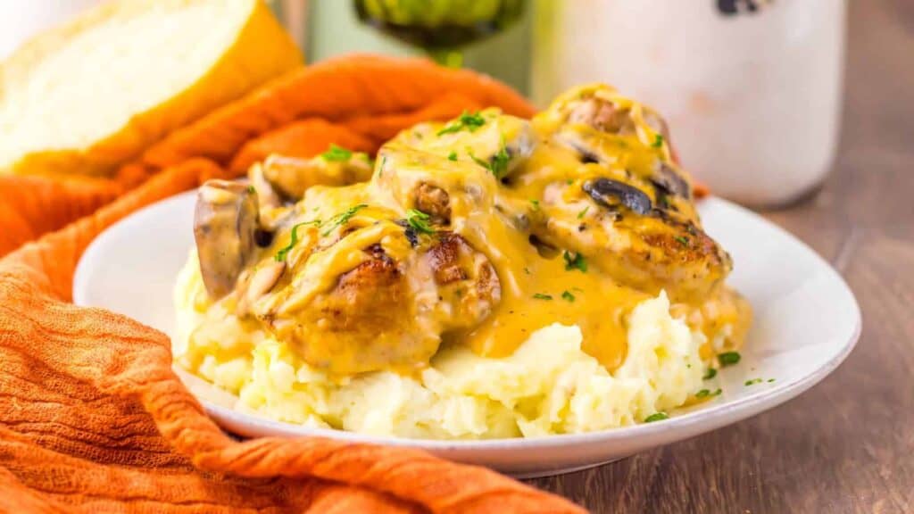 A plate of mashed potatoes topped with meatballs, sliced mushrooms, and creamy brown gravy, garnished with chopped parsley.