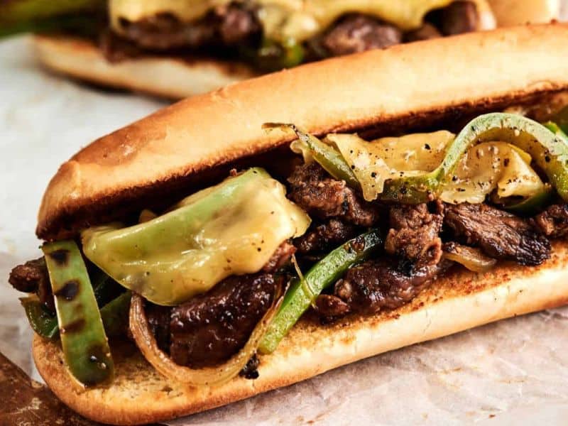 A sandwich roll filled with grilled steak strips, melted cheese, green bell peppers, and onions on parchment paper.