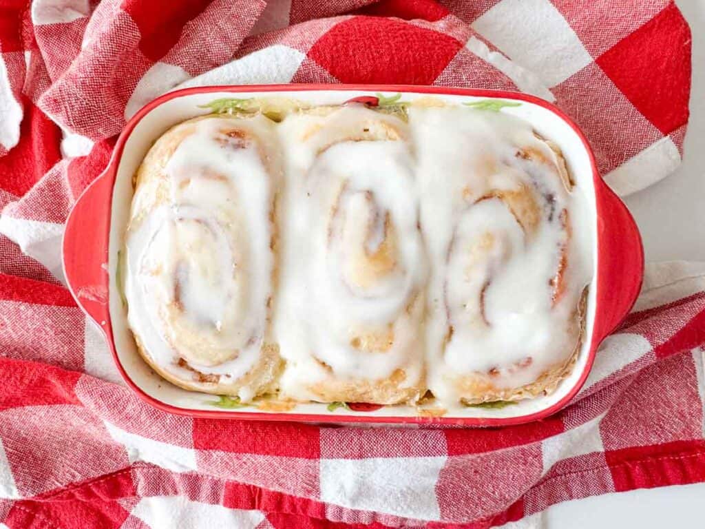 Three iced cinnamon rolls in a red baking dish sit on a red and white checkered cloth.