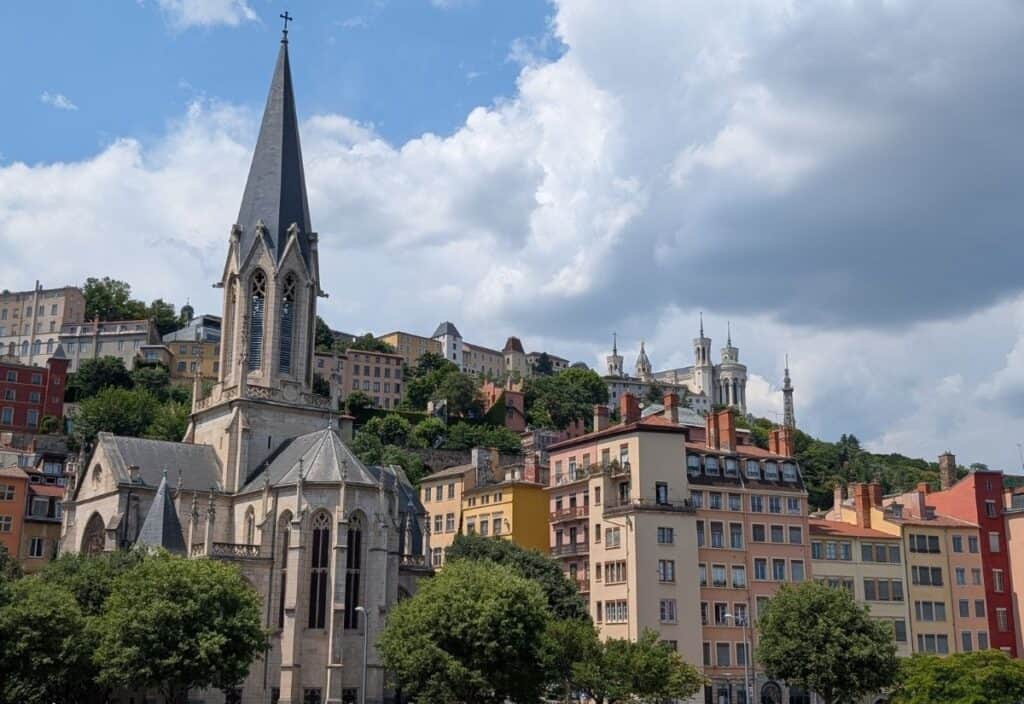 A stone church with a tall steeple stands before colorful buildings and a hill—an iconic scene for anyone spending 48 hours in Lyon, with more structures and trees visible against a partly cloudy sky.