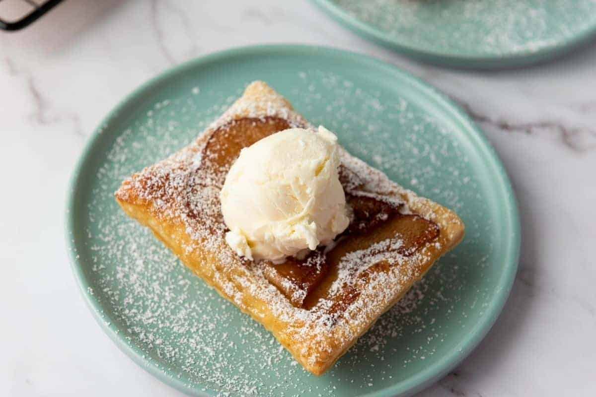 A Puff Pastry Apple Tart topped with a scoop of vanilla ice cream and sprinkled with powdered sugar sits on a light green plate.