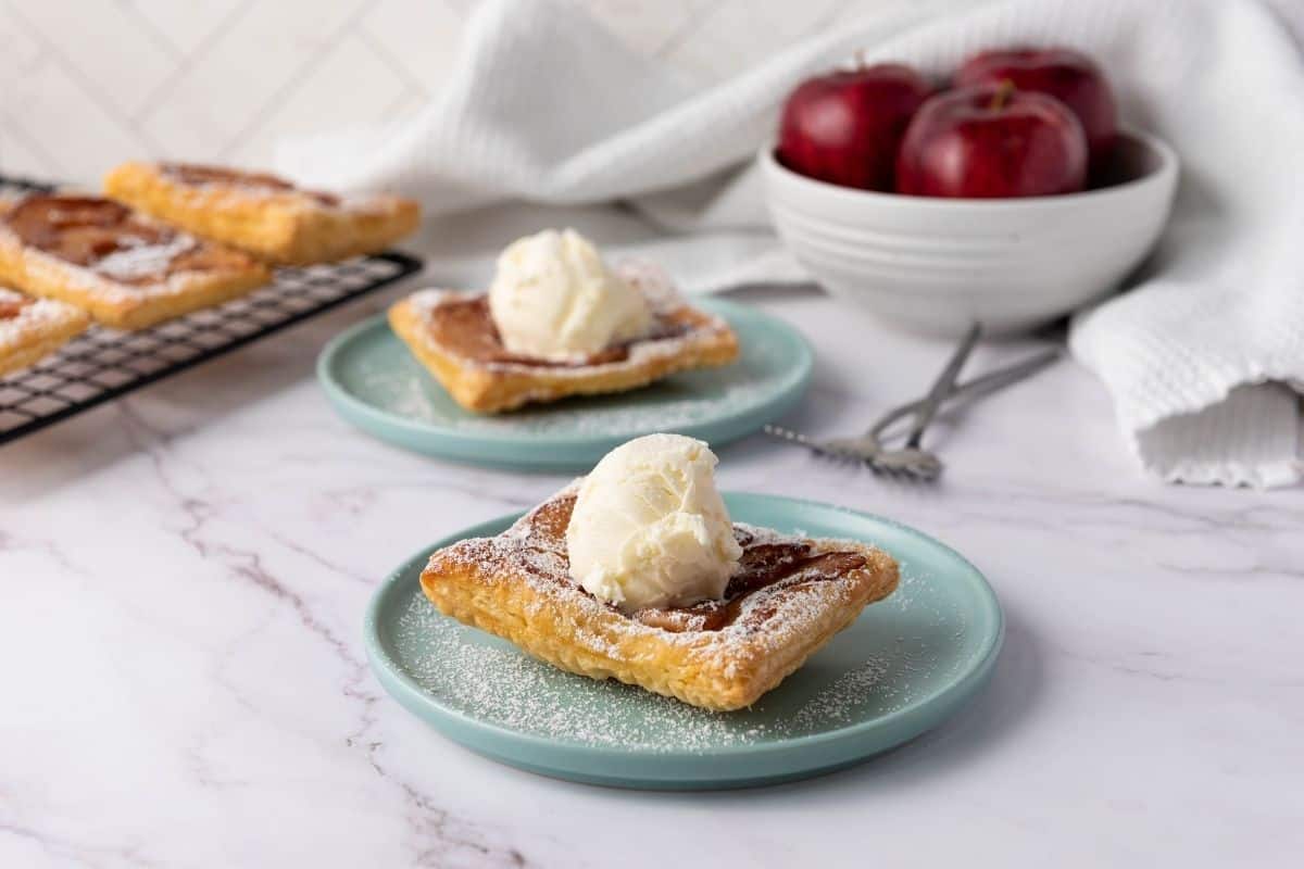 Two square apple pastries topped with vanilla ice cream are served on blue plates, with a bowl of red apples and a cooling rack with more pastries in the background.