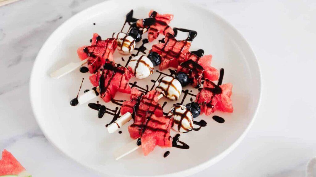 A white plate with skewers of watermelon, blueberries, and mozzarella balls, drizzled with balsamic glaze.