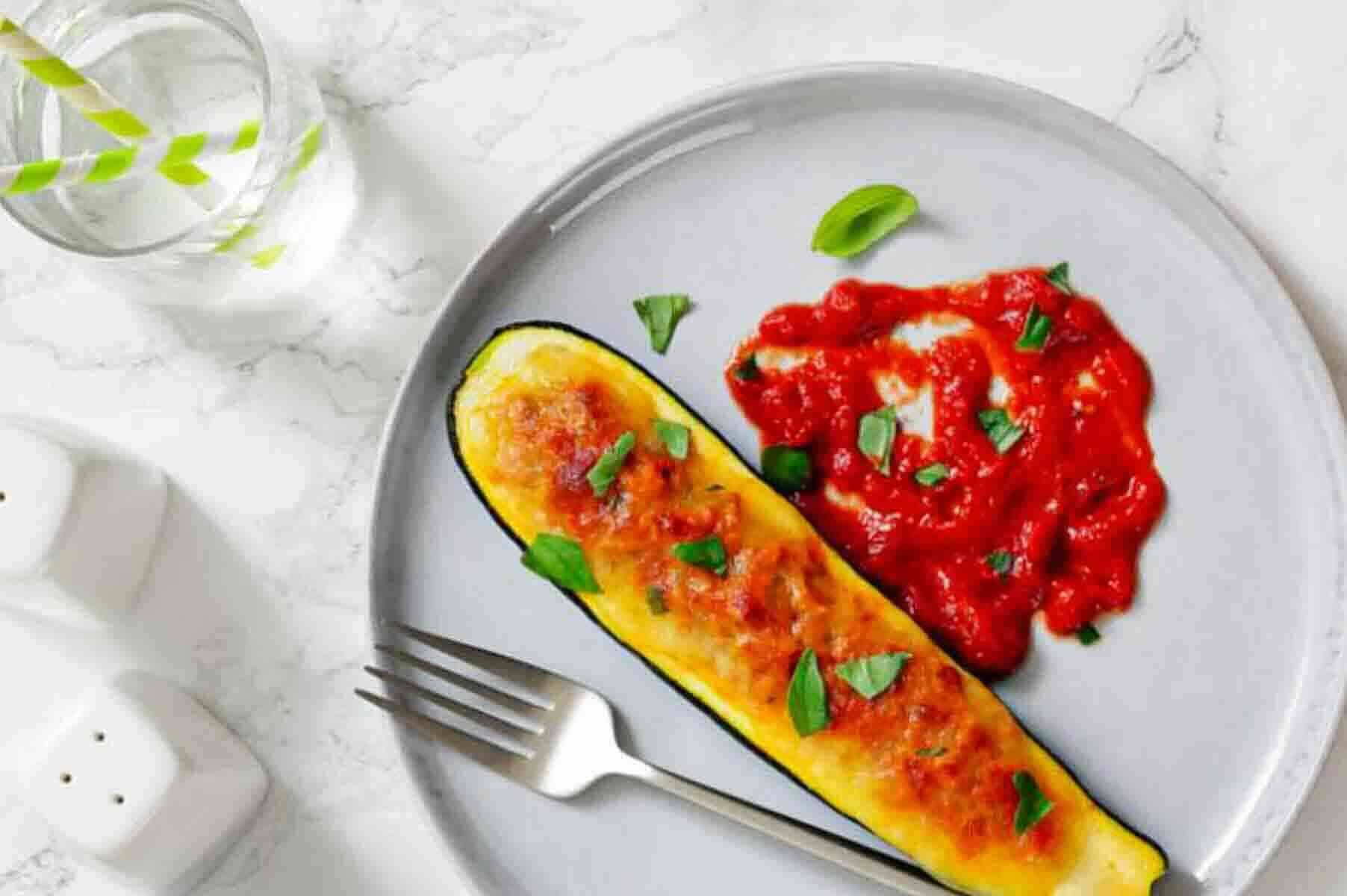 A stuffed zucchini boat topped with melted cheese and herbs, served with a portion of sauce on a gray plate, next to a fork and a glass of water.