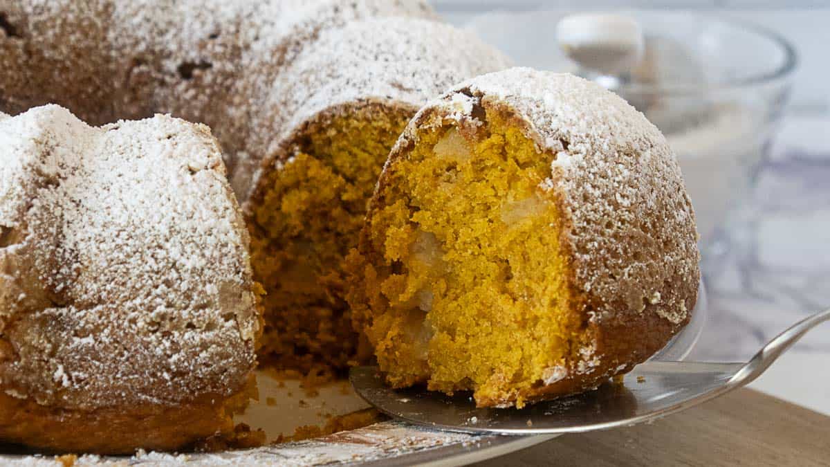 A slice of bundt cake with a golden, moist interior and chunks of fruit is being served; the cake is topped with powdered sugar.