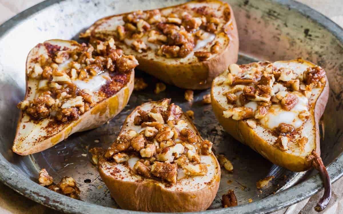 Halved pears in a metal baking dish, topped with chopped pecans, cinnamon, and melted butter or sauce.