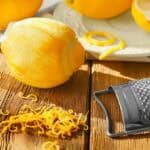 A peeled lemon, lemon zest, a grater, and whole lemons are arranged on a wooden surface with a plate in the background.