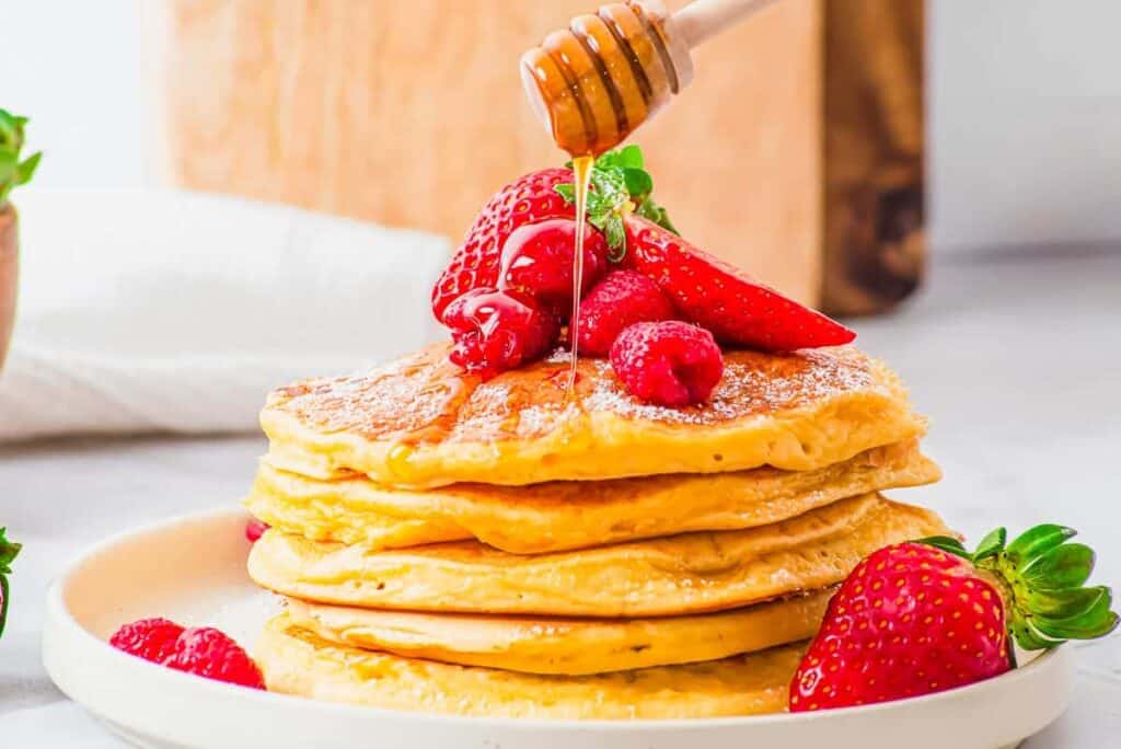 A stack of pancakes topped with strawberries and raspberries is being drizzled with honey on a white plate.