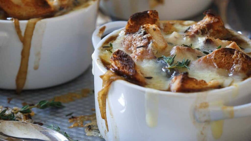 White ramekins filled with French onion soup, topped with toasted bread, melted cheese, and garnished with fresh thyme. Soup has slightly overflowed the sides.