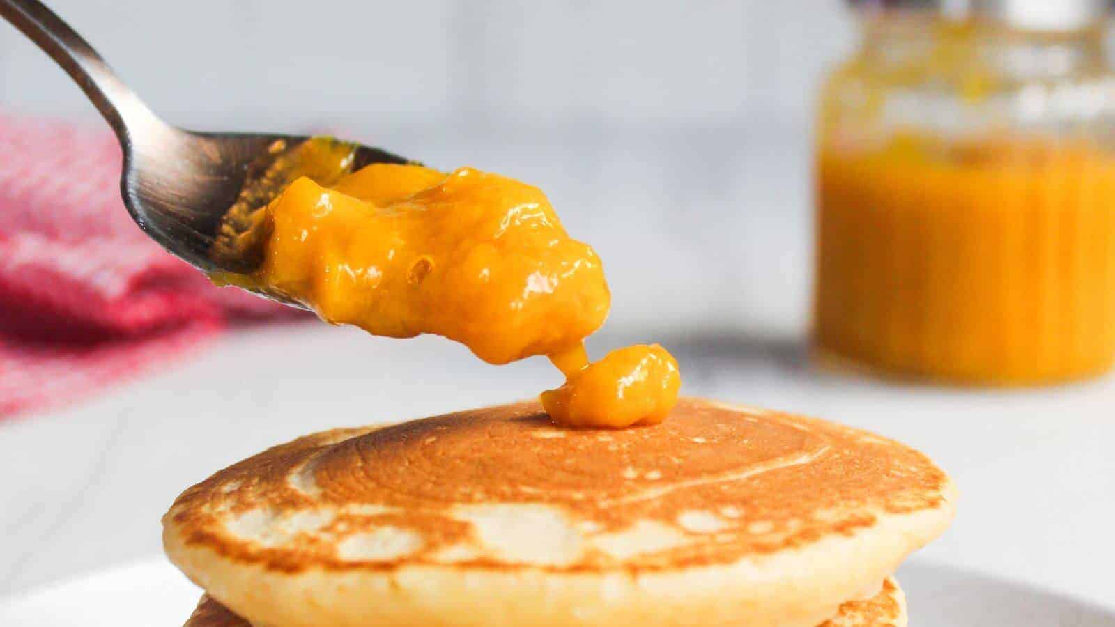 A spoonful of orange sauce is held above a stack of pancakes, with a jar of the same sauce blurred in the background.