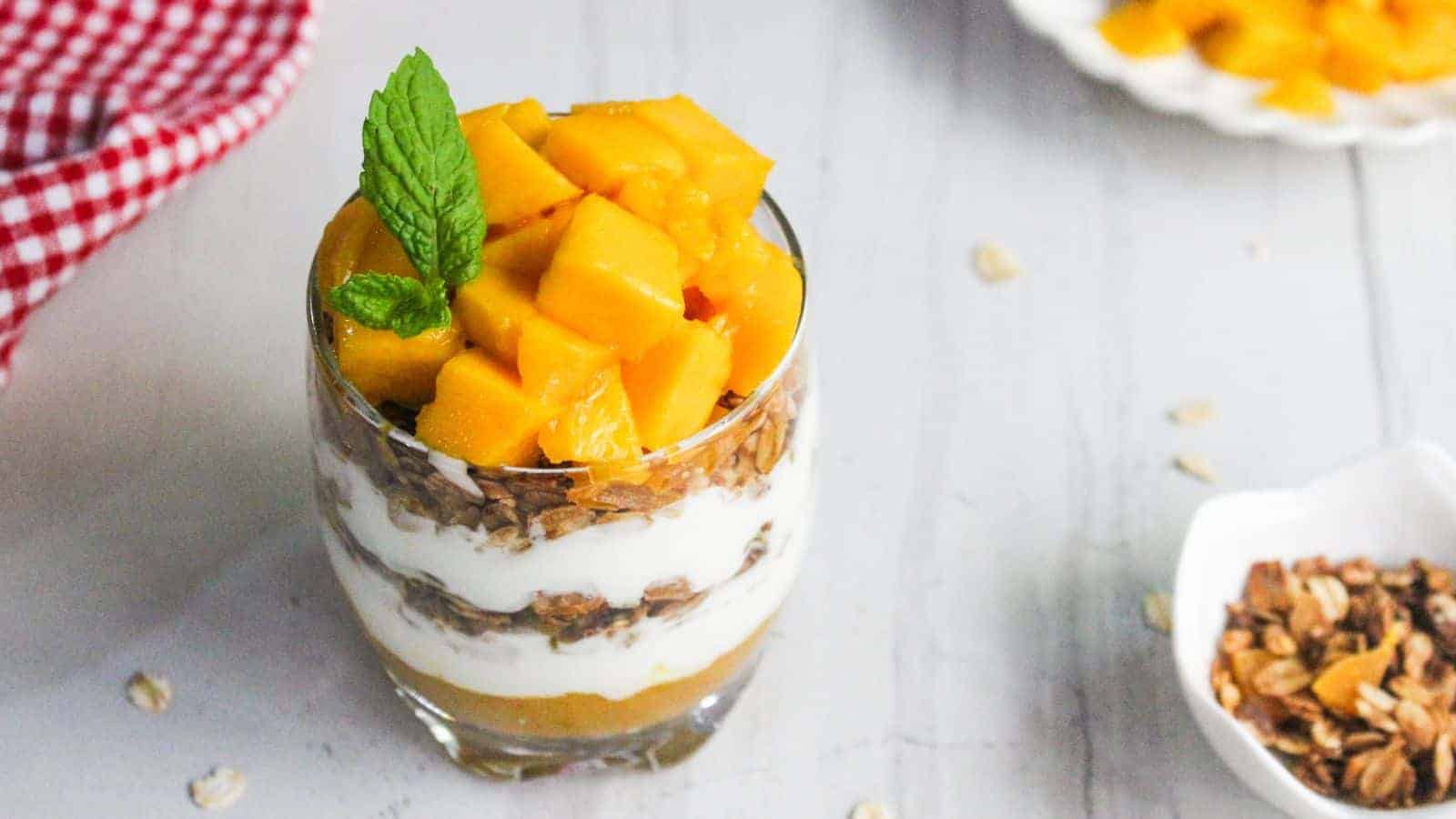 A glass filled with layers of yogurt, granola, and mango chunks, garnished with a mint leaf, placed on a light surface with scattered granola and oats nearby.