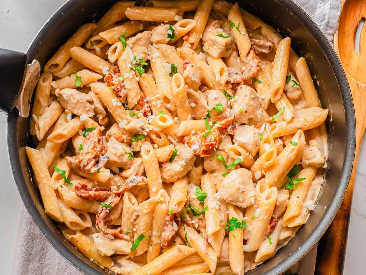 A skillet filled with penne pasta, pieces of chicken, sun-dried tomatoes, and a creamy sauce, garnished with chopped parsley.