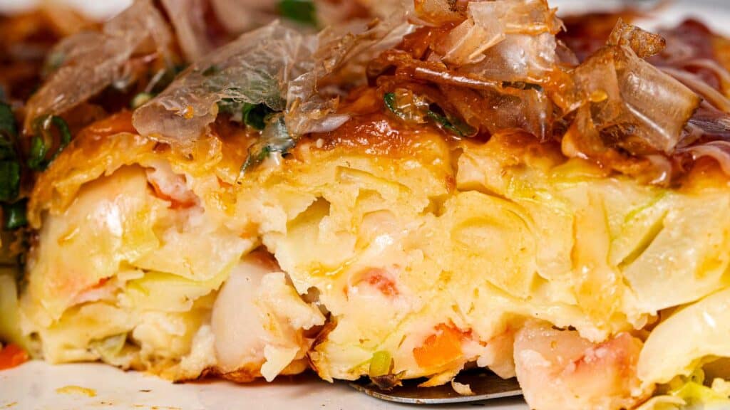 Close-up of a sliced okonomiyaki, showing its golden-brown crust, chopped vegetables, shrimp, and garnished with bonito flakes.