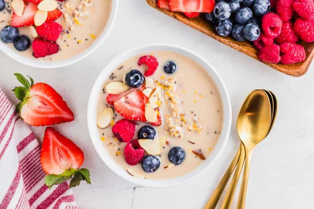 Bowl of smoothie topped with sliced strawberries, blueberries, raspberries, almond slivers, and chia seeds, next to gold spoons and a tray of fresh berries.
