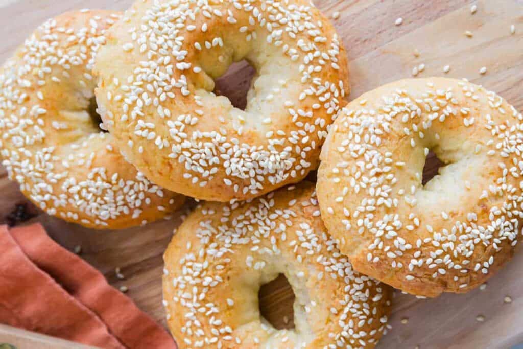 Four sesame seed bagels are arranged on a wooden surface with a rust-colored napkin partially visible at the corner.