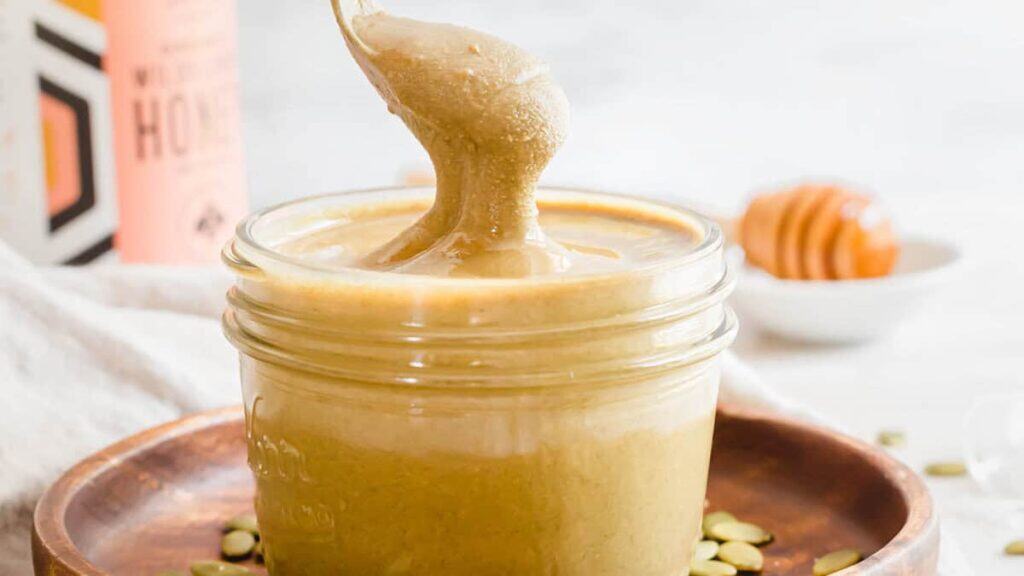 A spoon lifting creamy nut butter from a glass jar, with honey, a honey dipper, and a bottle in the background.