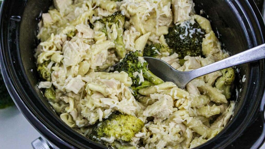 A slow cooker filled with cooked pasta, broccoli florets, shredded chicken, and creamy sauce, with a metal spoon resting inside.