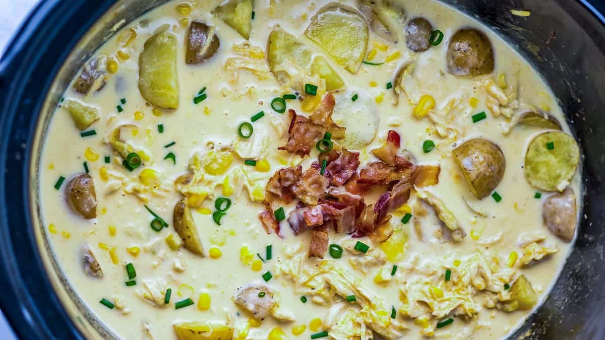 Creamy soup in a pot containing potatoes, corn, shredded chicken, chopped bacon, and garnished with chives.