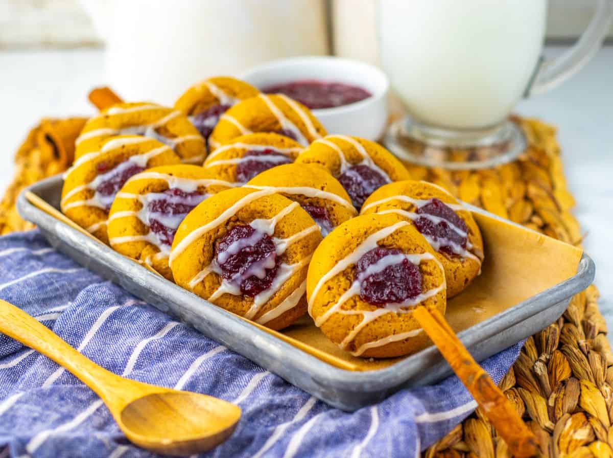 A tray of round cookies topped with white icing and a dollop of red jam, placed on parchment paper next to a bowl of jam and a glass of milk.