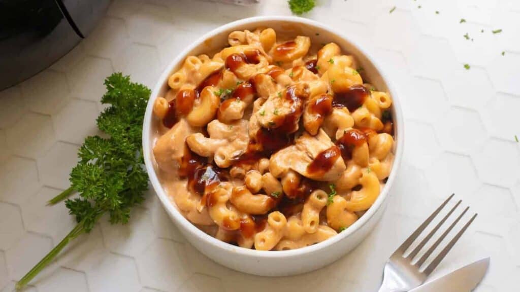 A bowl of macaroni and cheese with pieces of chicken, topped with barbecue sauce, placed on a white surface with parsley and a fork nearby.