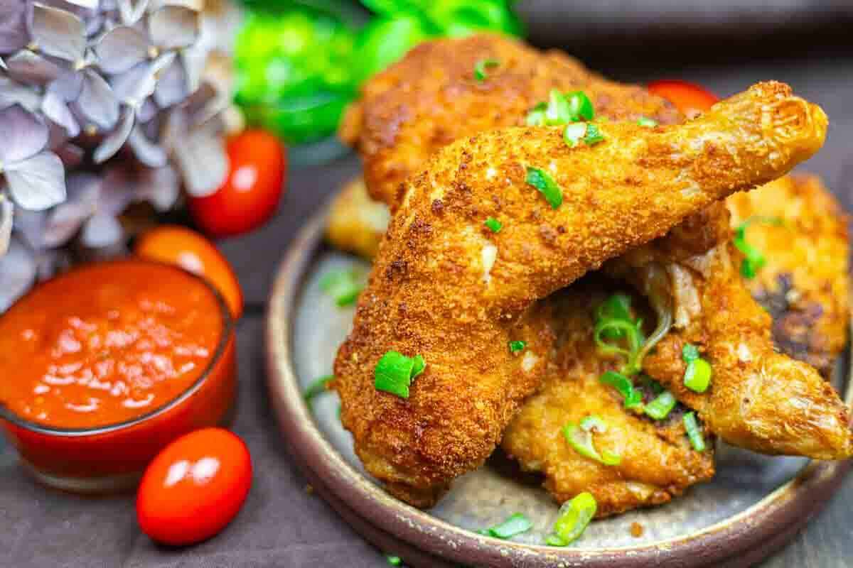 A plate of crispy fried chicken garnished with chopped green onions, served with a side of red dipping sauce and cherry tomatoes.