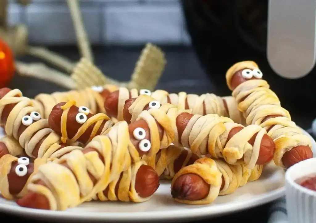 Hot dogs wrapped in crescent dough to resemble mummies, with candy eyes, arranged on a white plate.