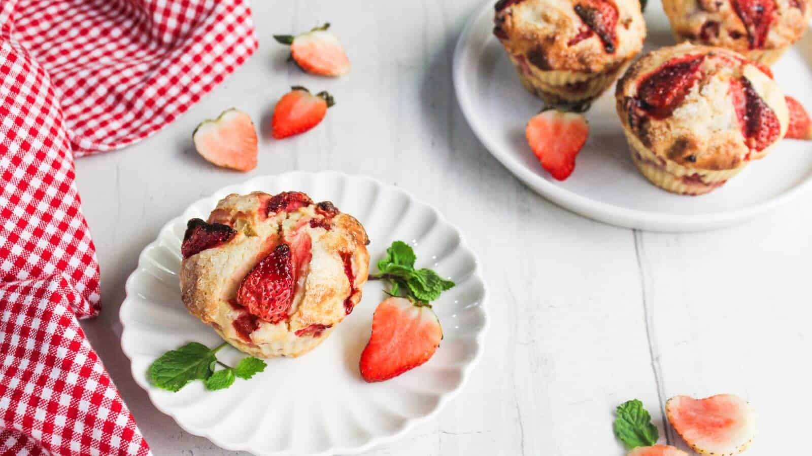 A strawberry muffin on a white plate with mint leaves and sliced strawberries, next to a red checkered cloth and more muffins on a separate plate.