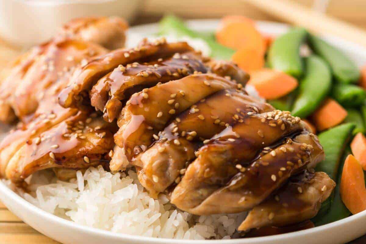 Plate of sliced teriyaki chicken over white rice, topped with sesame seeds, served with snap peas and carrot slices.