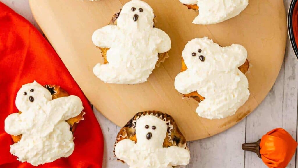 Six cupcakes decorated with white frosting shaped like ghosts are arranged on a wooden cutting board, with a red cloth and a small pumpkin nearby.