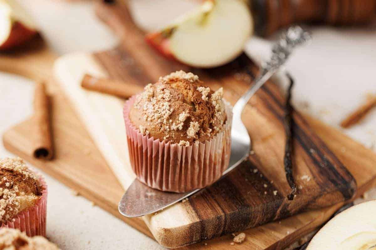 A muffin with crumb topping sits on a metal spatula over a wooden cutting board, surrounded by apple slices, cinnamon sticks, and a vanilla bean.