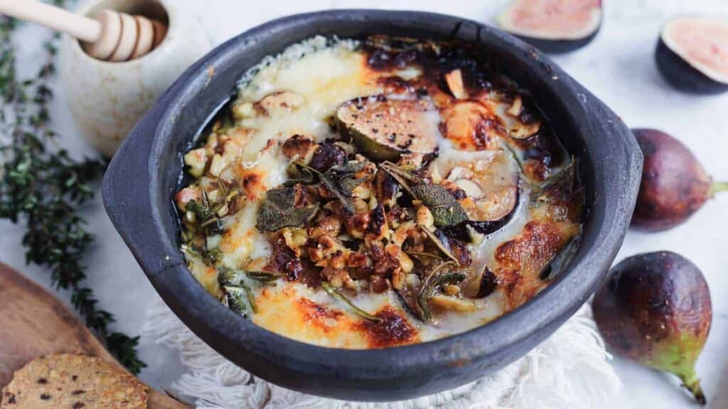 A baked cheese dish topped with figs, nuts, and herbs in a black ceramic bowl, with fresh figs and honey dipper in the background.