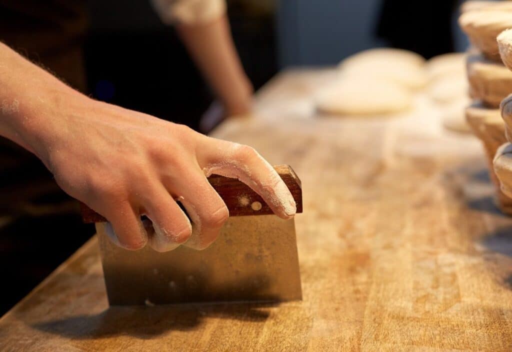A hand holding a metal bench scraper presses flour-dusted dough on a wooden surface, with stacks of dough portions in the background.
